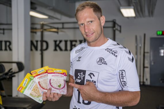 Ein Mann in einem weißen Sporttrikot steht in einer Turnhalle und zeigt auf drei Pakete mit Reissorten: Parboiled, Basmati und Langkorn, während er in die Kamera lächelt.