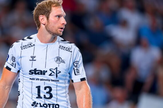 Ein männlicher Handballspieler in einem weißen Trikot mit schwarzen Akzenten und der Nummer 13 steht auf dem Spielfeld und schaut nach links. Im Hintergrund ist ein unscharfes Publikum zu sehen.