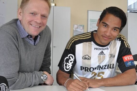 Zwei Männer posieren an einem Tisch; der eine lächelt in einer grauen Jacke, während der andere, der ein schwarz-goldenes Sporttrikot trägt, ein Dokument unterzeichnet. Im Hintergrund sind ein Schrank und eine Trophäe zu sehen.