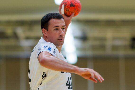 Ein männlicher Handballspieler in einem weißen Trikot bereitet sich darauf vor, einen orangefarbenen Ball während eines Spiels zu werfen, wobei er den Arm hebt und den Blick nach vorne richtet. Der Hintergrund des Hallenplatzes ist unscharf.