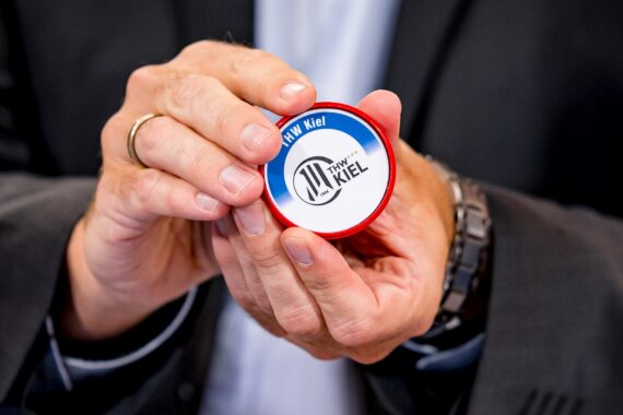 Eine Person im Anzug hält ein rundes Objekt mit blauem und rotem Rand und dem Logo des THW Kiel in der Hand, das einen Handball und einen stilisierten Text zeigt.