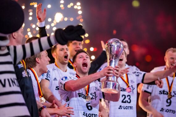 Eine jubelnde Handballmannschaft in weißen Trikots feiert, während ein Spieler eine Trophäe hochhält, umgeben von Mannschaftskameraden mit Medaillen und Konfetti, mit hellen Funken im Hintergrund.