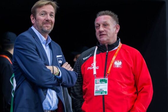Zwei Männer stehen nebeneinander; der eine trägt einen marineblauen Blazer und ein hellblaues Hemd, der andere eine rote Sportjacke mit dem polnischen Wappen und einem Akkreditierungsband. Sie scheinen bei einer Hallensportveranstaltung zu sein.