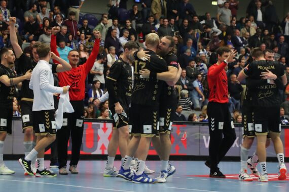 Eine Handballmannschaft in schwarz-goldenen Trikots feiert auf dem Spielfeld, umarmt sich und jubelt, während im Hintergrund eine Menge Zuschauer zuschaut und klatscht.