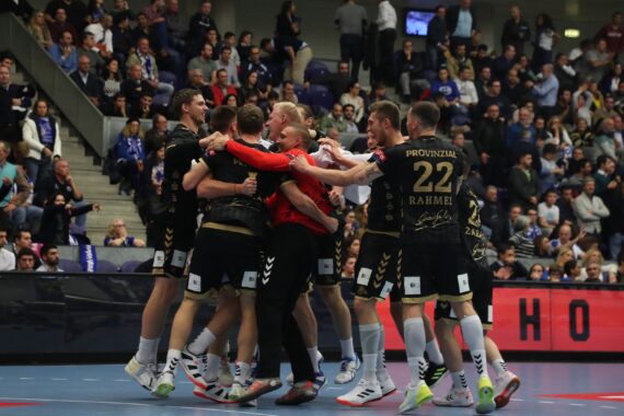 Eine Gruppe von Handballspielern in schwarzen Trikots feiert gemeinsam auf einem Hallenplatz, umarmt sich und lächelt, während eine große Menge von Zuschauern von der Tribüne im Hintergrund zusieht.