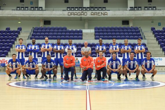 Ein Herrensportteam in blau-weißen Uniformen stellt sich für ein Gruppenfoto auf einem Hallenplatz in der Dragão-Arena auf. Davor knien drei Trainer in orangefarbenen Jacken und im Hintergrund sind leere blaue Sitze zu sehen.