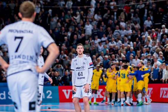 Eine Szene aus einem Handballspiel zeigt zwei Spieler in weißen Trikots, die einer Gruppe von Spielern in Gelb gegenüberstehen und feiern. Die Zuschauer auf der Tribüne schauen im Hintergrund aufmerksam zu.