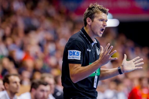 Ein Handballtrainer in einem schwarzen Hemd steht an der Seitenlinie und gibt mit offenen Händen leidenschaftlich Anweisungen, während im Hintergrund eine verschwommene Menge und Spieler zu sehen sind.