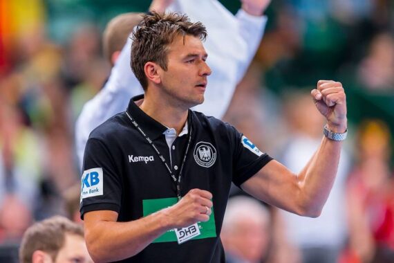 Ein Mann, der ein schwarzes Polohemd mit Aufnähern und einem Schlüsselband trägt, hebt seine Faust in einer Geste des Jubels oder der Ermutigung während einer Sportveranstaltung. Der Hintergrund ist unscharf und zeigt eine Sporthalle.