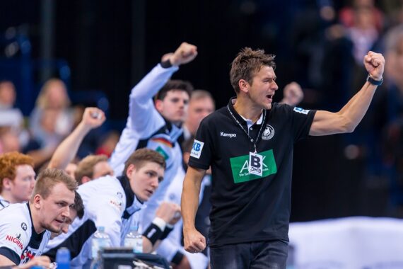 Ein Handballtrainer in einem schwarzen Hemd jubelt leidenschaftlich mit seiner Mannschaft, die während eines Spiels auf der Bank ebenfalls die Fäuste zum Jubel hebt. Der Hintergrund zeigt eine Sportarena mit unscharfen Zuschauern.