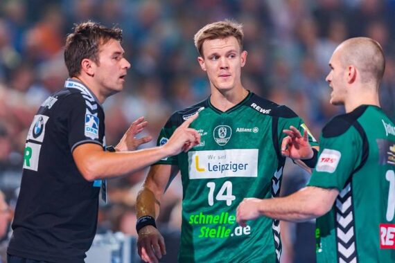 Drei männliche Handballspieler in grün-schwarzen Trikots diskutieren während eines Spiels auf dem Spielfeld, während im Hintergrund eine unscharfe Menschenmenge zu sehen ist. Der Spieler in der Mitte trägt die Nummer 14.