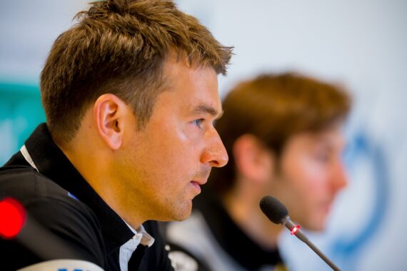 Ein Mann in einem schwarzen Hemd spricht auf einer Pressekonferenz in ein Mikrofon, während eine andere Person im Hintergrund verschwommen zu sehen ist. Der Fokus liegt auf dem Gesicht des Mannes im Profil.