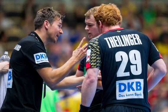 Ein Trainer gestikuliert und spricht intensiv mit zwei Handballspielern auf dem Spielfeld. Die Spieler tragen schwarze Trikots, einer trägt die THEILINGER 29 auf dem Rücken. Die Szene spielt sich während eines Spiels ab, mit unscharfen Zuschauern im Hintergrund.