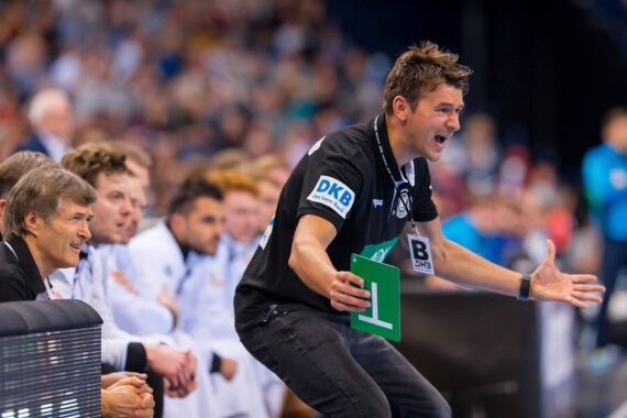 Ein leidenschaftlicher Handballtrainer gestikuliert energisch von der Seitenlinie aus und hält ein grünes Schild in die Höhe, während Spieler und Betreuer im Hintergrund aufmerksam das Spiel verfolgen.