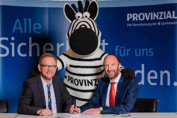 Zwei Männer in Anzügen sitzen an einem Tisch und unterschreiben Dokumente, während sie in die Kamera lächeln. Hinter ihnen steht eine Person in einem Zebra-Maskottchenkostüm mit der Aufschrift PROVINZIAL auf der Brust. Im Hintergrund ist eine blaue Fahne mit deutschem Text zu sehen.