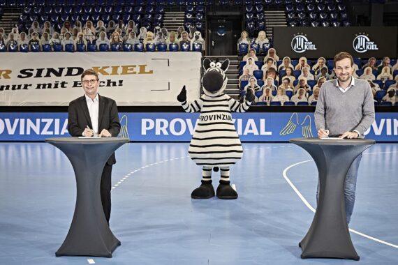 Zwei Männer stehen auf einem Sportplatz an verschiedenen Podien und unterschreiben Dokumente. Eine Person in einem Zebra-Maskottchenkostüm steht zwischen ihnen und zeigt den Daumen nach oben. Pappfiguren von Menschen füllen die Plätze im Hintergrund.