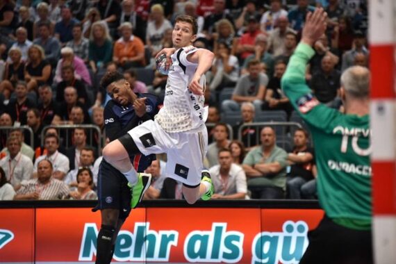 Ein Handballspieler in einem weißen Trikot versucht, den Ball zu schießen, während ein Verteidiger in Blau versucht, ihn zu blocken. Ein Torwart in Grün bereitet sich darauf vor, den Schuss zu stoppen. Das Publikum im Hintergrund schaut aufmerksam zu.