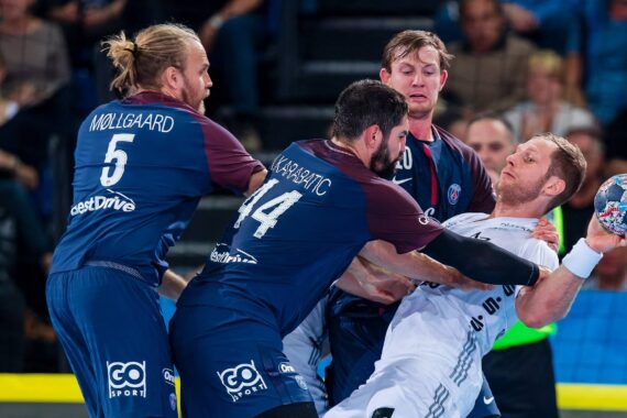 Drei Handballspieler in blauen Trikots verteidigen sich gegen einen Spieler in einem weißen Trikot, der den Ball hält, während sie sich während eines Spiels heftig streiten, während die Zuschauer im Hintergrund zusehen.