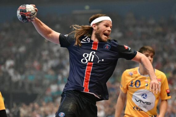 Ein männlicher Handballspieler in einem dunkelblauen Trikot springt mit einem Ball in der Hand und bereitet sich auf den Wurf vor, während ein Gegner in einem gelben Trikot zusieht. Die Arena ist voller Zuschauer im Hintergrund.
