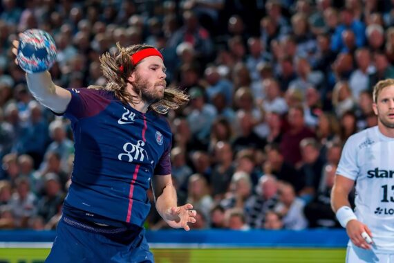 Ein Handballspieler in einer marineblauen Uniform mit rotem Stirnband bereitet sich auf den Wurf vor, während ein anderer Spieler in einer weißen Uniform zuschaut. Der Hintergrund zeigt eine unscharfe Menschenmenge in einem Stadion.