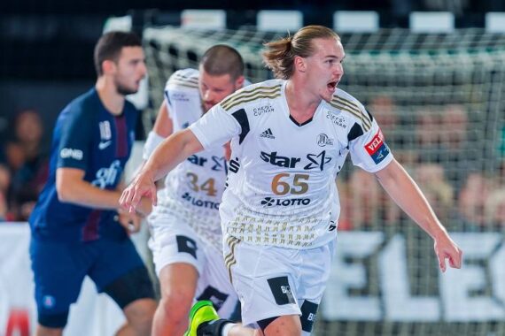 Ein Handballspieler in einem weißen Trikot mit der Nummer 65 jubelt auf dem Spielfeld, während im Hintergrund in der Nähe des Tores Mannschaftskameraden und ein Gegner zu sehen sind.