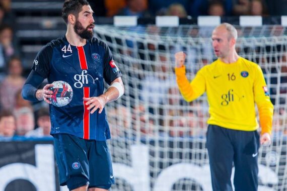 Ein männlicher Handballspieler in einem dunkelblauen PSG-Trikot hält den Ball auf dem Spielfeld, während ein Mannschaftskamerad in einem gelben Torwarttrikot im Hintergrund in der Nähe des Tors die Faust hebt.