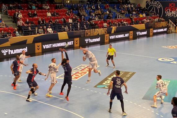 Handballspieler in blau-weißen Trikots liefern sich auf einem Hallenplatz ein intensives Spiel, bei dem ein Spieler den Ball abblockt oder abfängt. Die Zuschauer schauen von der Tribüne aus zu, und Banner säumen die Arena.
