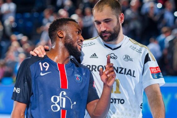 Zwei männliche Handballspieler, einer in einem dunkelblauen Trikot (Nummer 19) und der andere in einem weißen Trikot, stehen nach einem Spiel dicht beieinander auf dem Spielfeld, unterhalten sich und lächeln, während die Zuschauer im Hintergrund verschwommen zu erkennen sind.