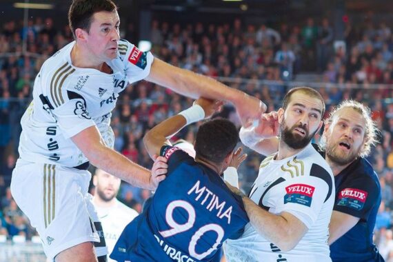 Eine Gruppe von männlichen Handballspielern ist während eines Spiels in Aktion. Ein Spieler in Weiß springt, um den Ball zu werfen, während andere in blauen und weißen Trikots eng um ihn herum spielen. Die Zuschauer sind im Hintergrund unscharf zu erkennen.