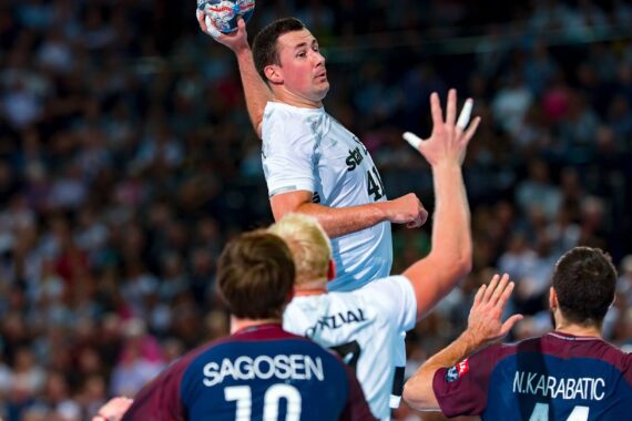 Ein Handballspieler in einem weißen Trikot springt, um den Ball zu schießen, umgeben von Verteidigern in kastanienbraunen Trikots, während eines intensiven Spiels in einer überfüllten Arena.