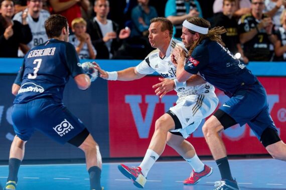 Drei männliche Handballspieler liefern sich einen intensiven Wettkampf; der eine in Weiß versucht, mit dem Ball vorzudringen, während zwei in Blau ihn abwehren, während im Hintergrund eine Menschenmenge zusieht.