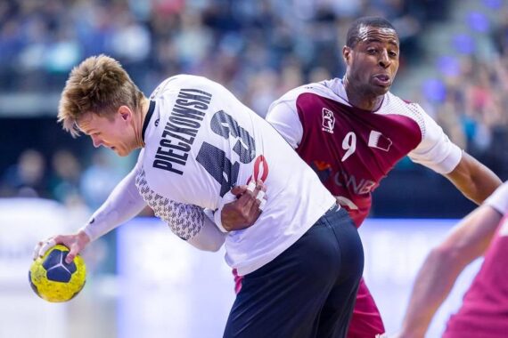 Zwei männliche Handballspieler in Aktion auf dem Spielfeld; der eine trägt ein weißes Trikot mit der Aufschrift PIECZKOWSKI 19 und hält den Ball, während der andere in einer kastanienbraunen Uniform versucht, ihn zu verteidigen. Im Hintergrund ist ein unscharfes Publikum zu sehen.