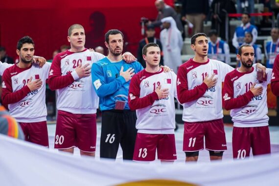 Eine Gruppe männlicher Handballspieler in kastanienbraunen und weißen Uniformen steht in einer Reihe mit der rechten Hand über dem Herzen und singt vor einem Spiel. Im Vordergrund ist eine große Flagge zu sehen.