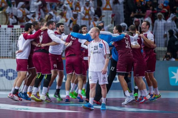 Ein Handballspieler in einem weißen Trikot steht im Vordergrund, während im Hintergrund eine Mannschaft in kastanienbraunen Trikots gemeinsam feiert, während die Zuschauer von der Tribüne aus zusehen.