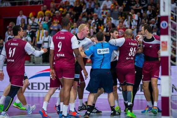 Handballspieler gegnerischer Mannschaften stehen sich auf dem Spielfeld gegenüber, während die Schiedsrichter versuchen, sie zu trennen, während die Zuschauer im Hintergrund zuschauen. Einige Spieler tragen weiße, andere kastanienbraune Katar-Trikots.