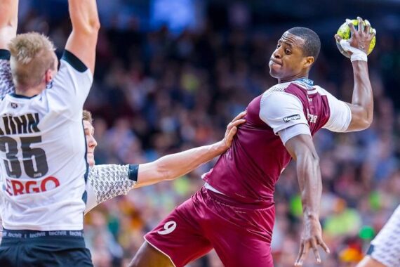 Ein Handballspieler in einem kastanienbraunen Trikot springt, um den Ball zu werfen, während er während eines Spiels von zwei Spielern in weißen und schwarzen Trikots verteidigt wird. Die Aktion ist intensiv, und die Zuschauer sind im Hintergrund verschwommen.