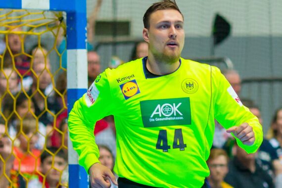 Ein männlicher Handballtorwart in einem leuchtend gelben Trikot mit der Nummer 44 steht während eines Spiels in der Nähe des Torpfostens, während im Hintergrund Zuschauer zu sehen sind.