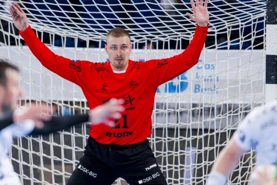 Ein männlicher Handballtorwart in einem roten Trikot steht mit erhobenen Armen, bereit, das Tor zu verteidigen. Im Vordergrund sind Spieler in weißen Trikots zu sehen, und hinter ihm ist das Netz zu sehen.