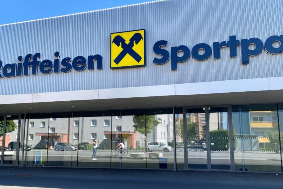 Das Bild zeigt die Außenseite eines Gebäudes mit dem Zeichen Raiffeisen Sportpark und einem gelben Logo. Das Glas spiegelt nahe gelegene Gebäude und eine vorbeigehende Person.