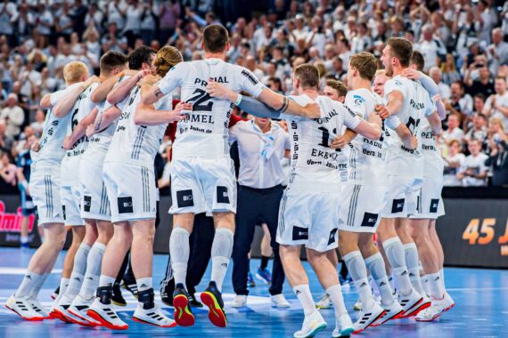 Eine Männer-Handballmannschaft in weißen Trikots steht in einer Halle und springt zusammen. Die Zuschauer im Hintergrund jubeln und sind überwiegend in Weiß gekleidet.