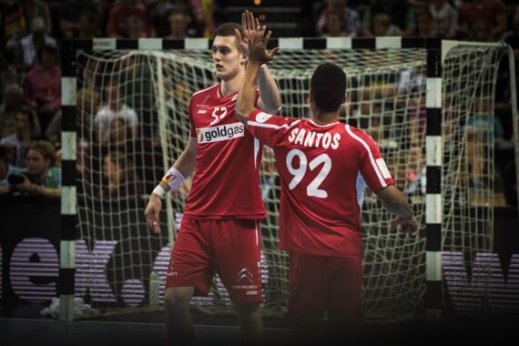 Zwei männliche Handballspieler in roten Trikots klatschen sich während eines Spiels vor einem Tor ab, wobei das Trikot des einen Spielers den Namen "Santos" und die Nummer 92 zeigt. Im Hintergrund sind Zuschauer zu sehen.