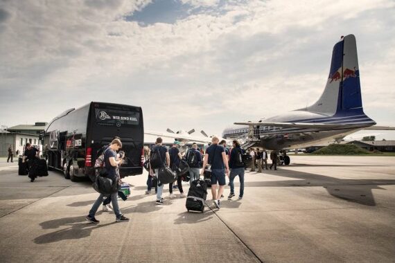Eine Gruppe von Menschen mit Gepäck geht aus einem Bus in Richtung eines Oldtimer-Flugzeugs auf dem Rollfeld eines Flughafens unter einem teilweise bewölkten Himmel.
