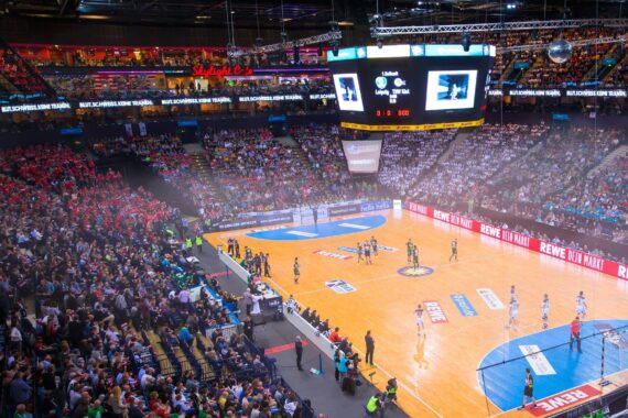 Eine große, mit Fans gefüllte Halle, die ein Handballspiel verfolgen. Die Spieler sind auf dem Spielfeld, und die Anzeigetafel zeigt den Spielstand an. Die Zuschauer füllen die Sitze, mit Anfeuerungsrufen auf beiden Seiten. Helle Lichter erhellen die Szene.