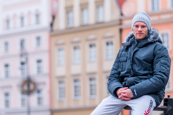 Eine Person, die eine dunkle Winterjacke, eine graue Mütze und eine helle Jogginghose trägt, sitzt im Freien mit gefalteten Händen vor einem Hintergrund aus pastellfarbenen Gebäuden.