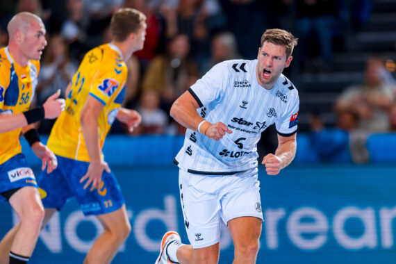 Ein männlicher Handballspieler in einem weißen Trikot jubelt während eines Spiels mit einer geballten Faust, während sich Spieler in gelben Trikots im Hintergrund auf dem Spielfeld bewegen.