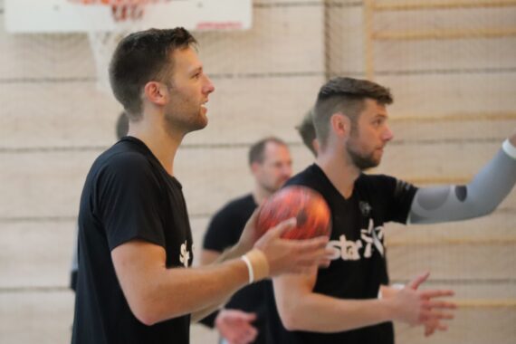 Zwei Männer in schwarzen Hemden halten einen Basketball in der Hand und reichen ihn weiter. Ein Mann bereitet sich darauf vor, den Ball zu werfen, während der andere seinen Arm hebt. Im Hintergrund sind ein Basketballkorb und unscharfe Spieler zu sehen.