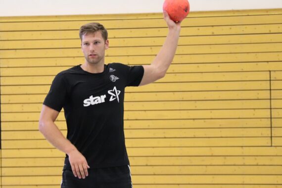 Ein Mann in schwarzem T-Shirt und Shorts steht in einem Haus und hält mit einer Hand einen roten Ball über dem Kopf, als wolle er ihn werfen. Im Hintergrund ist eine gelbe Holzwand zu sehen.