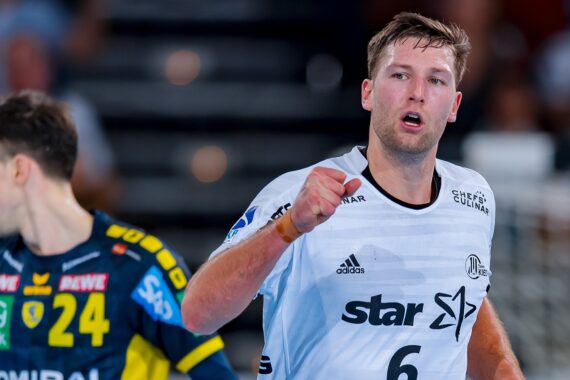 Ein männlicher Handballspieler in einem weißen Trikot mit der Nummer 6 jubelt mit geballter Faust, während ein anderer Spieler in einem blau-gelben Trikot mit der Nummer 24 in der Nähe steht. Der Hintergrund ist unscharf.