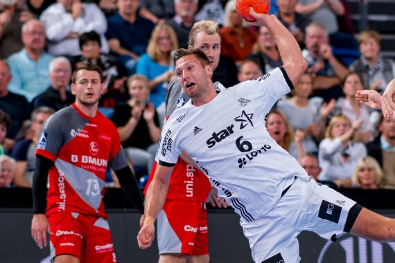 Ein Handballspieler in einem weißen Trikot wirft gerade einen orangefarbenen Ball und ist während eines Hallenspiels von gegnerischen Spielern in roten Trikots umgeben. Die Zuschauer im Hintergrund schauen gebannt zu.