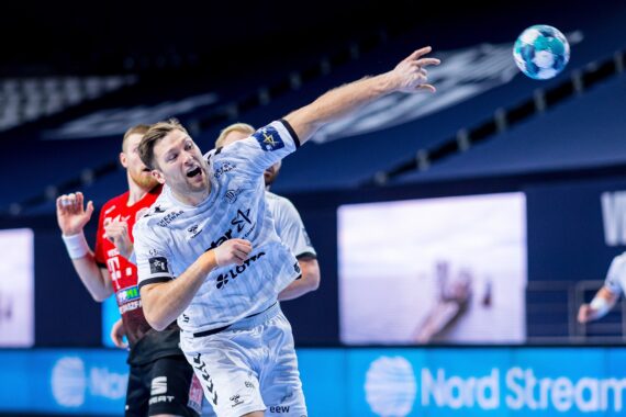 Ein männlicher Handballspieler in einem weißen Trikot wirft den Ball während eines Spiels in die Luft, während im Hintergrund andere Spieler und eine blaue Nord Stream-Werbung zu sehen sind.